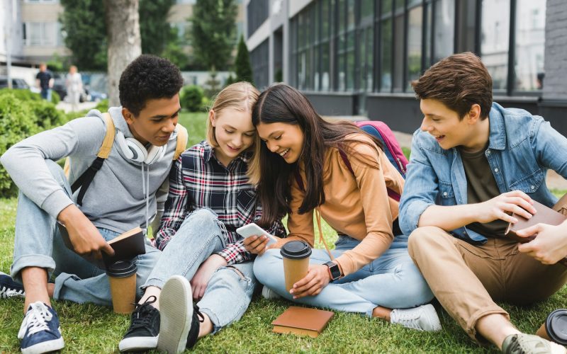 smiling-and-happy-teenagers-sitting-on-grass-and-looking-at-smartphone.jpg